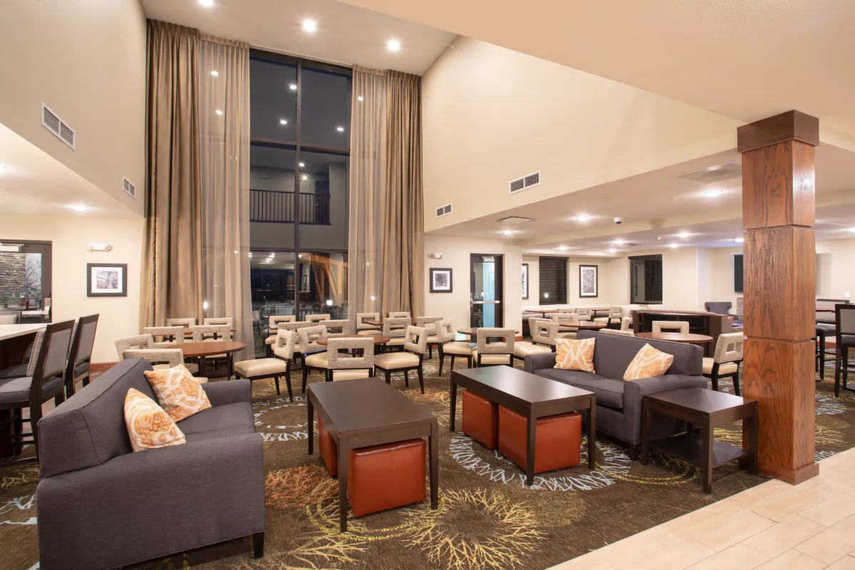 A warmly lit interior lounge area featuring large windows that flood the space with natural light. The area is furnished with plush, comfortable seating and chic tables, and accented by decorative curtains. The overall ambiance is cozy and inviting, perfect for relaxing, reading, or enjoying a drink.