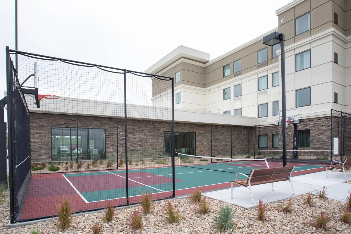 The image presents an energetic outdoor basketball court adjacent to the hotel, with benches and the building's facade in the background. People are shown engaged in a friendly game and others relaxing on benches, reflecting opportunities for recreation and informal networking.