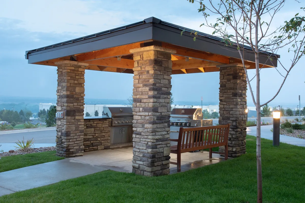 This exterior scene showcases a welcoming outdoor area with stone columns, grill stations, and a wooden bench. The space is designed for casual socializing, barbecuing at sunset, and relaxing under the stars, evoking a sense of community and connection to the outdoors.