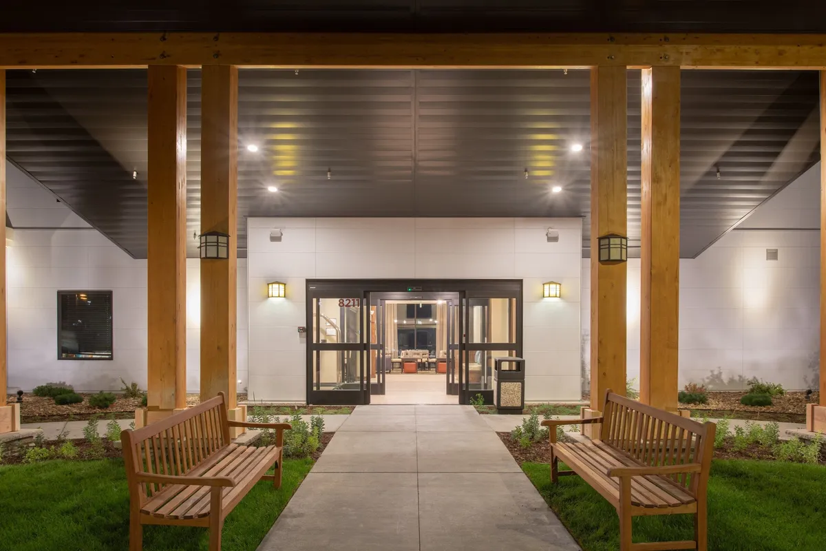 A warm, inviting view of the hotel's landscaped front entrance, complete with wooden benches and a lush garden. The setup encourages guests to gather, relax, or enjoy a welcoming atmosphere upon arrival or before heading out to nearby dining and entertainment options.